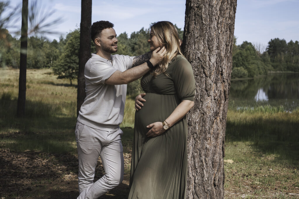 Stralende zwangere vrouw in een mooie groene plisse jurk die tegen een boom aanstaat, liefdevol naar haar partner kijkt die een haar uit haar gezicht strijkt